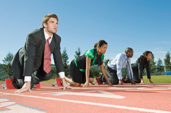 Vier Menschen stehen in Bürokleidung in Startposition auf einer Laufbahn.