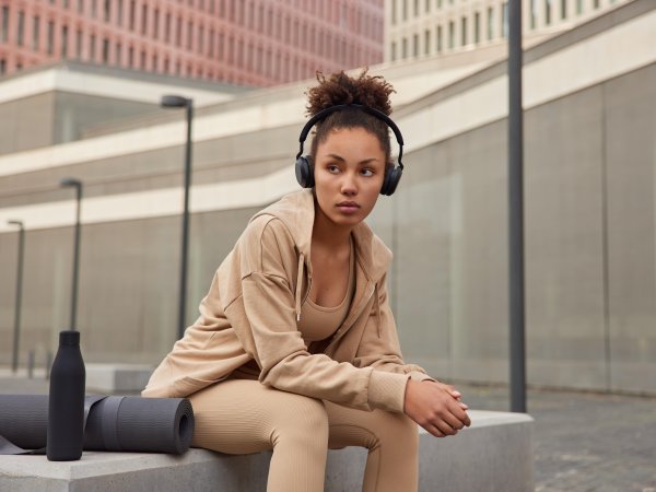 A woman sits on a bench in a city and listens to music through headphones. She is wearing light brown to beige athleisure wear.