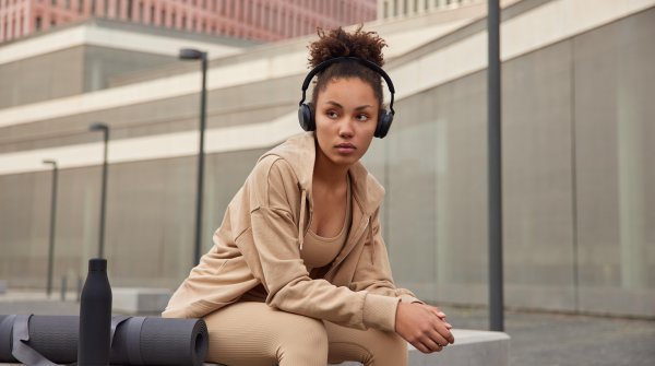 A woman sits on a bench in a city and listens to music through headphones. She is wearing light brown to beige athleisure wear.