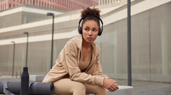 Eine Frau sitzt auf einer Bank in einer Stadt und hört Musik über Kopfhörer. Sie trägt hellbraune bis beige Athleisure Wear.