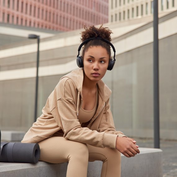 A woman sits on a bench in a city and listens to music through headphones. She is wearing light brown to beige athleisure wear.