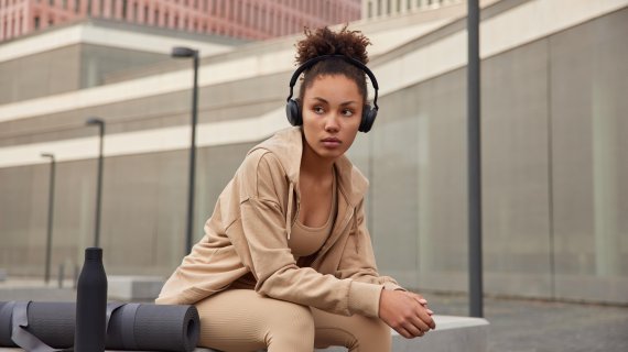 A woman sits on a bench in a city and listens to music through headphones. She is wearing light brown to beige athleisure wear.