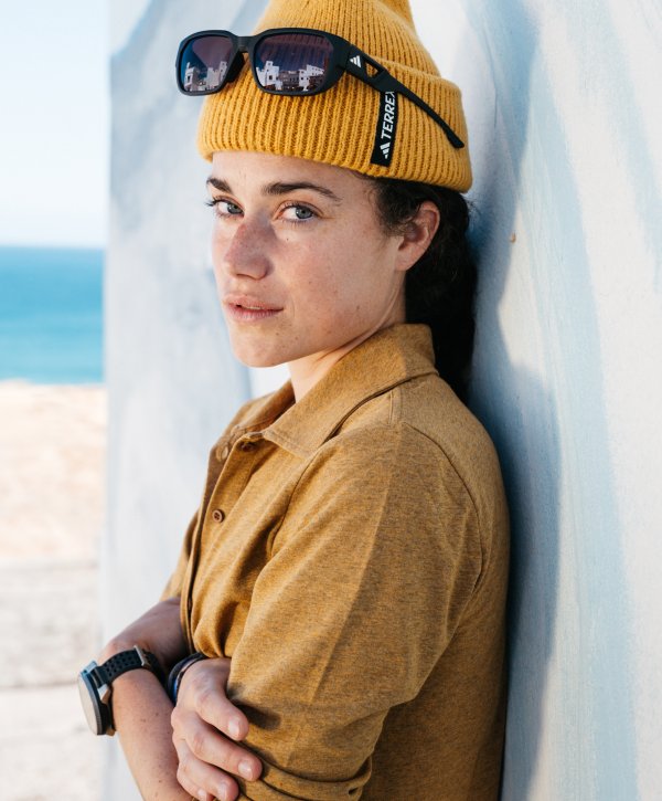 Trailsrunnerin Kimi Schreiber in gelber Kleidung an Wand gelehnt; Hintergrund: Meer und Strand